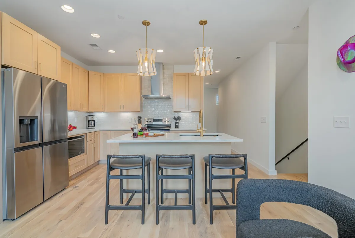 A bright, modern kitchen with a large island at its center, surrounded by bar stools. Stainless steel appliances line the back wall, illuminated by stylish pendant lights overhead. The space invites both cooking and entertaining, with ample counter space for food preparation or casual gatherings.
