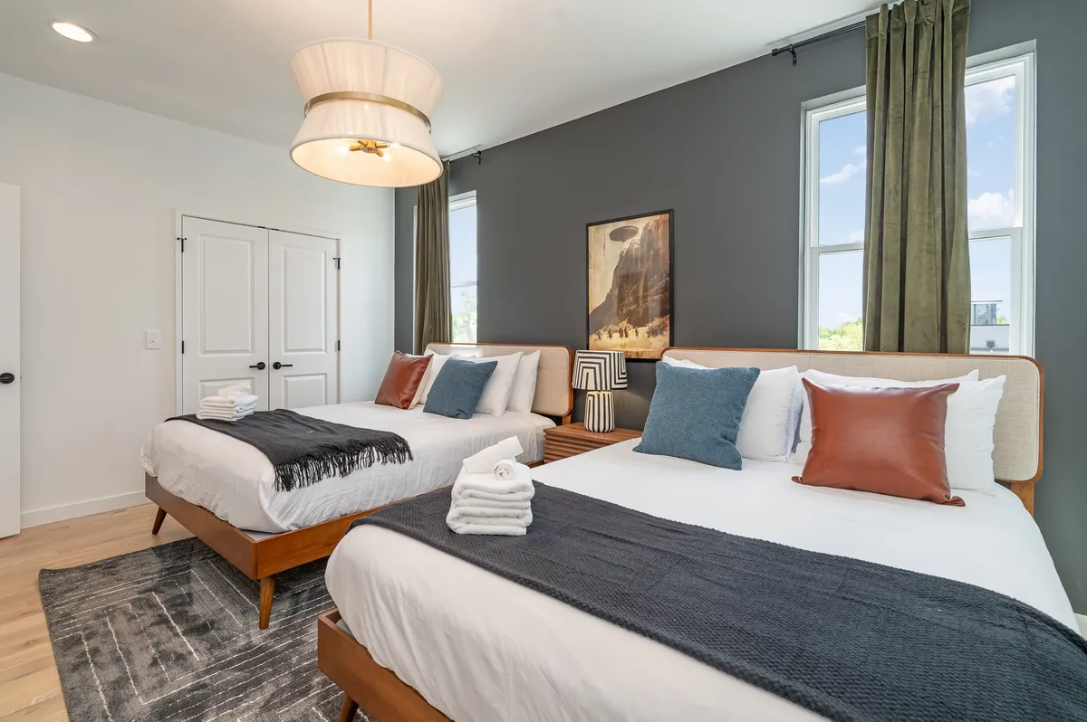 A bright, comfortable room with two double beds, large windows letting in natural light, stylish artwork, and a contemporary pendant light fixture. The setup is both inviting and flexible for groups needing extra sleeping arrangements.