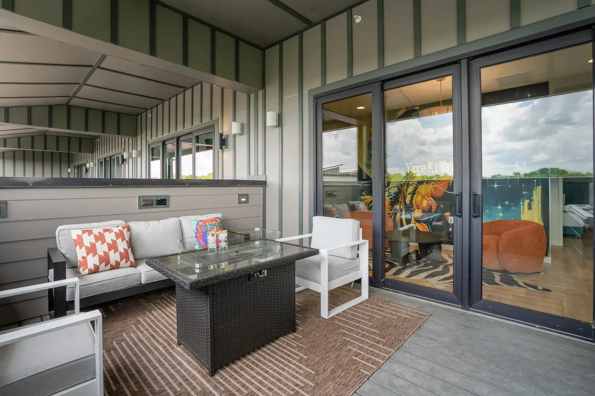 A stylish outdoor seating area on a rooftop deck, featuring a glass coffee table and colorful cushions. The area is illuminated by natural light, with panoramic city views in the background, creating a vibrant setting for morning coffee or sunset celebrations.