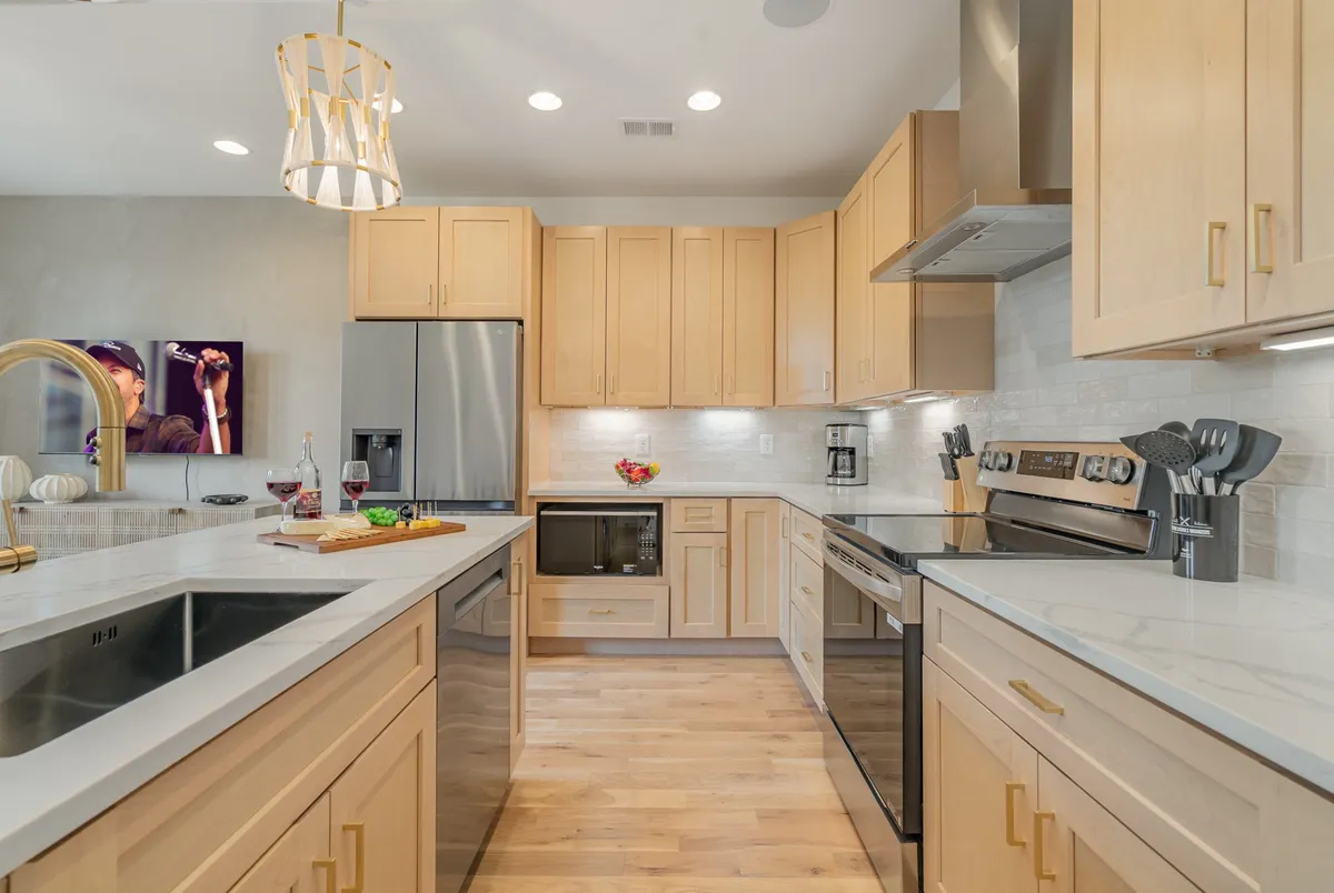 A welcoming kitchen scene with a prominent island, shiny stainless steel appliances, and a golden light fixture adding warmth. Fruits and wine are arranged on the counter, suggesting the space is ready for hosting and shared meals.